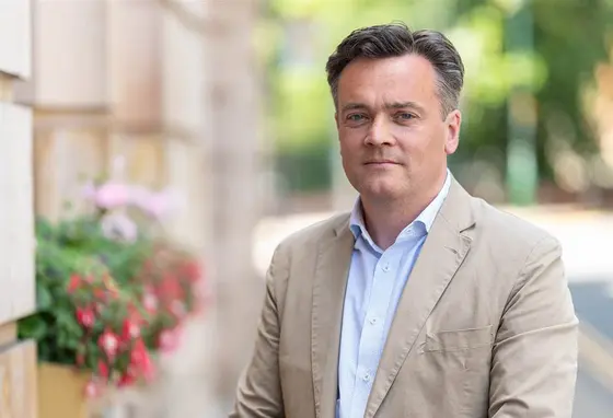 James Twining wearing shirt and jacket, standing next to a stone building with flower boxes.