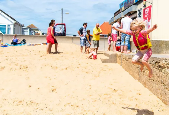 A young girl jumping from a wall onto the sand at Jaywick in Essex. Families are in the background at a cafe.