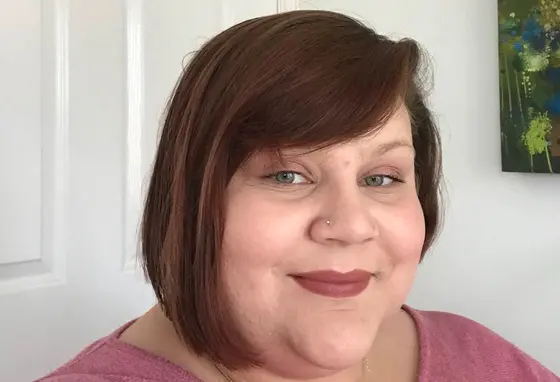 A selfie of a women with shoulder-length brown hair, standing against a white wall