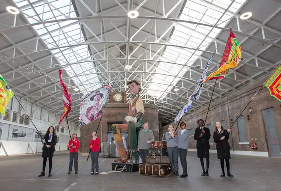 School children holding large flags stand in an empty station building accompanied by a giant puppet.