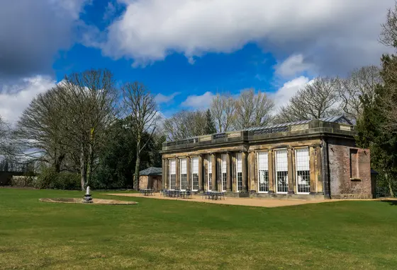 A Georgian stone building with glass roof and large windows. A lawn and stone fountain are in front of the building