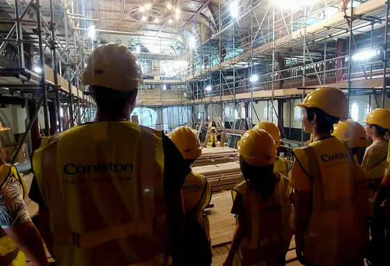 a group of people wearing hard hats and high-vis jackets stand inside the building looking at conservation works