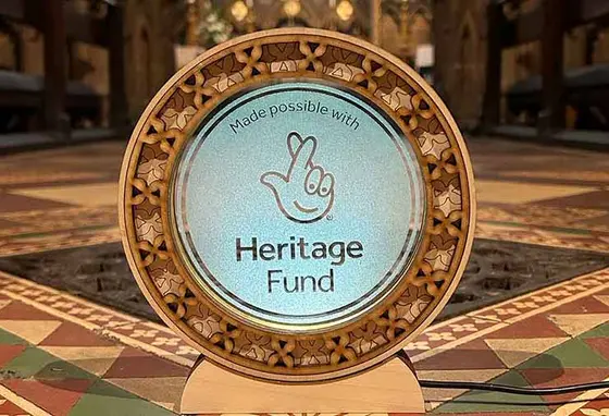 The National Lottery Heritage Fund acknowledgement stamp etched onto a circular glass plaque with a fretwork boarder. The plaque sits in front of a colourful tiled floor and an ironwork grate.