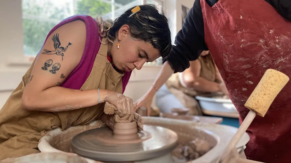 A person in an apron helping another person use a pottery wheel