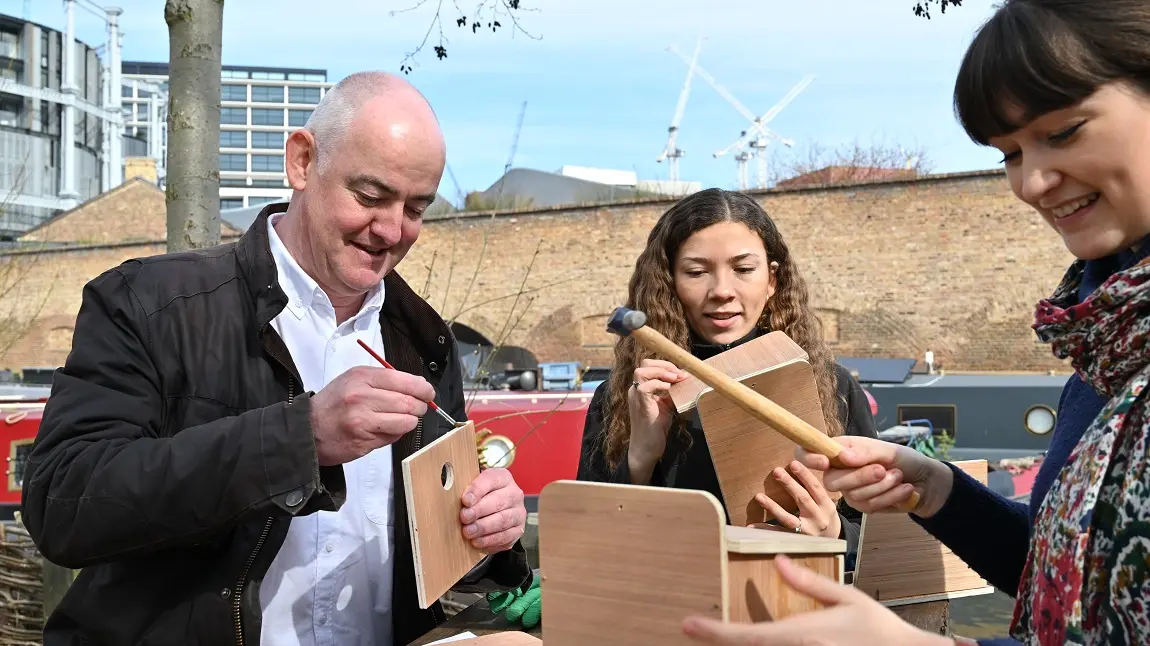 Three people sitting on a picnic table in an urban greenspace, building wooden birdboxes with hammers 