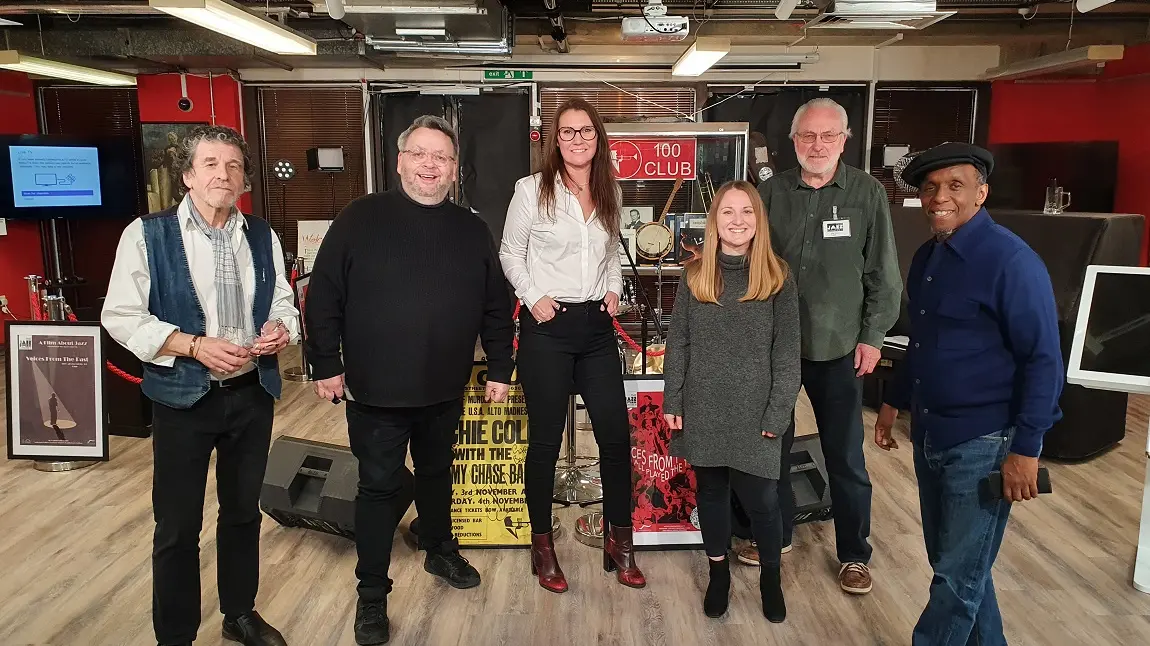 Six people posing for a picture in front of a Jazz music archive, including posters and frames 