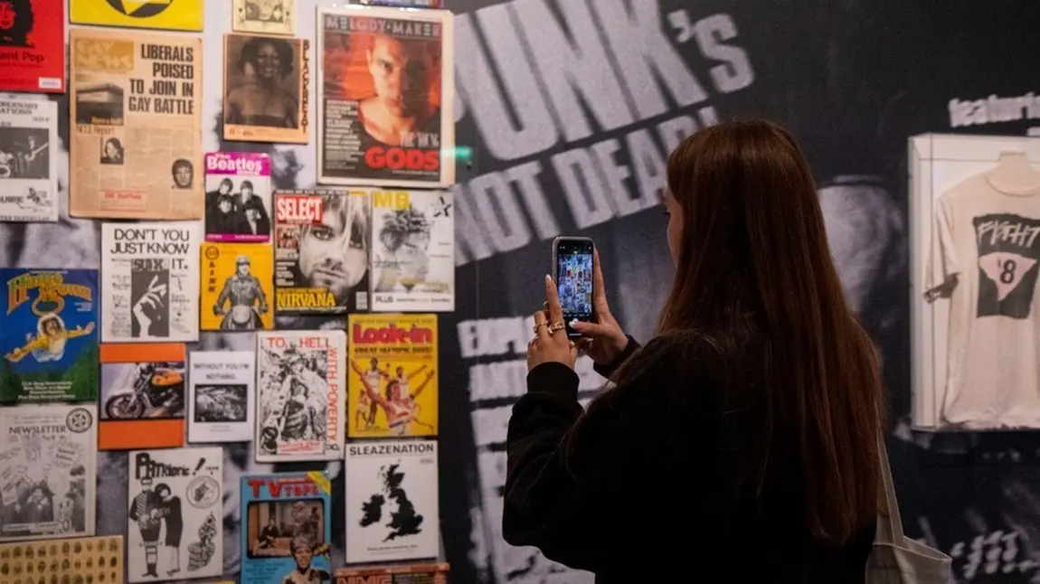 Image of person with long dark hair and dark clothing taking a picture on a mobile of a display of music gig posters at The Museum of Youth Culture