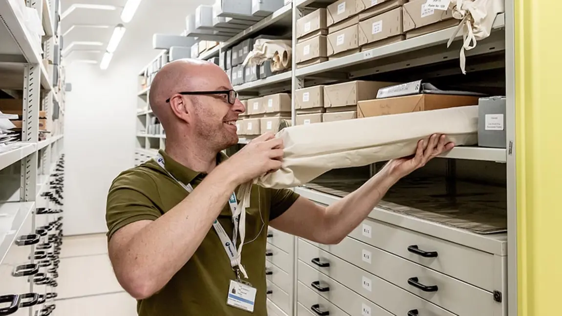 a man takes a cloth bag containing a rolled up document off a shelf in an archive