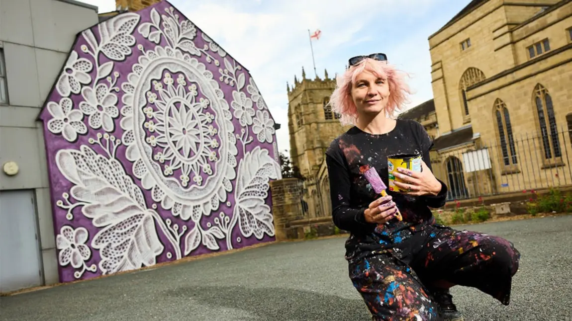 An artist kneels in front of a painted wall with an intricate pattern on a purple background