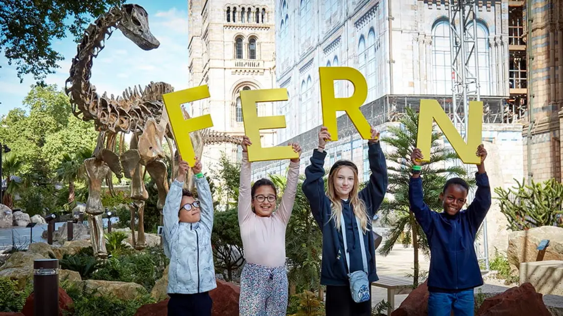 four children hold up large letters spelling FERN, the name of the diplodocus skeleton on display behind them