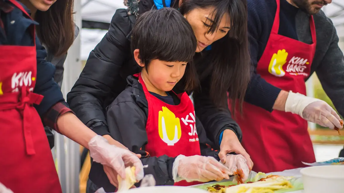 A child in an apron making kimchi with the help of an adult