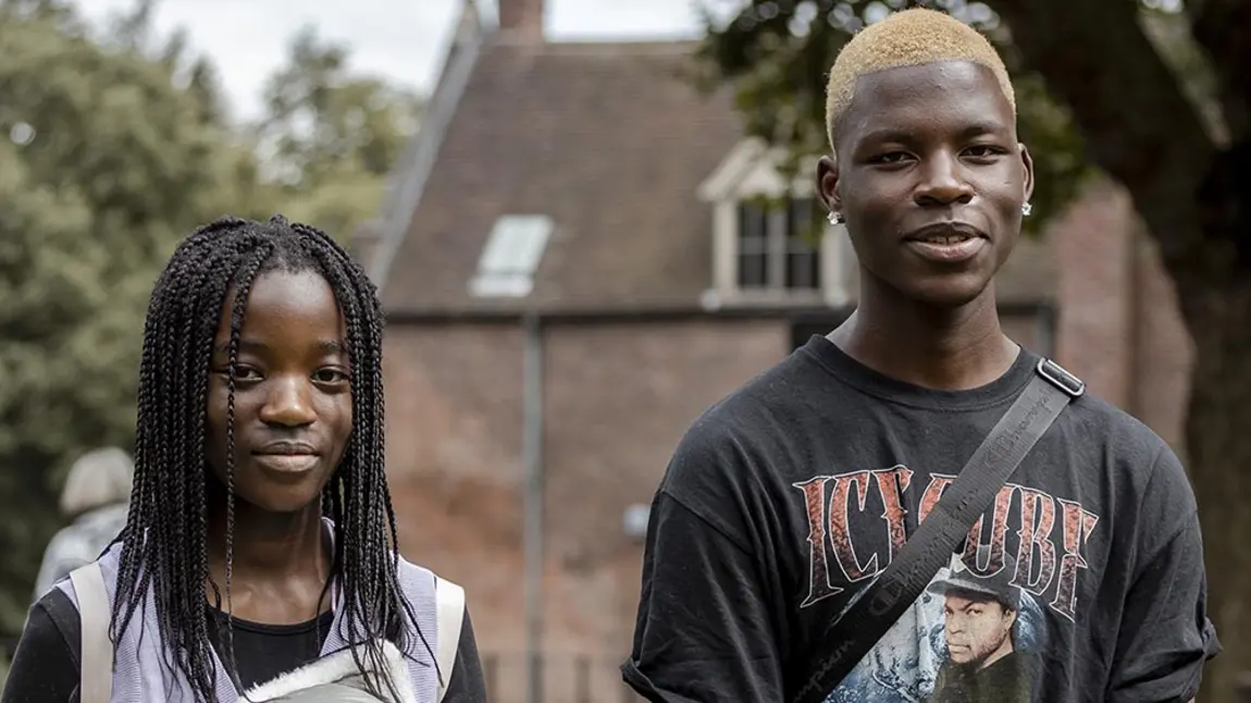 Two young black people wearing casual clothes stand outside with a historic brick building in the background