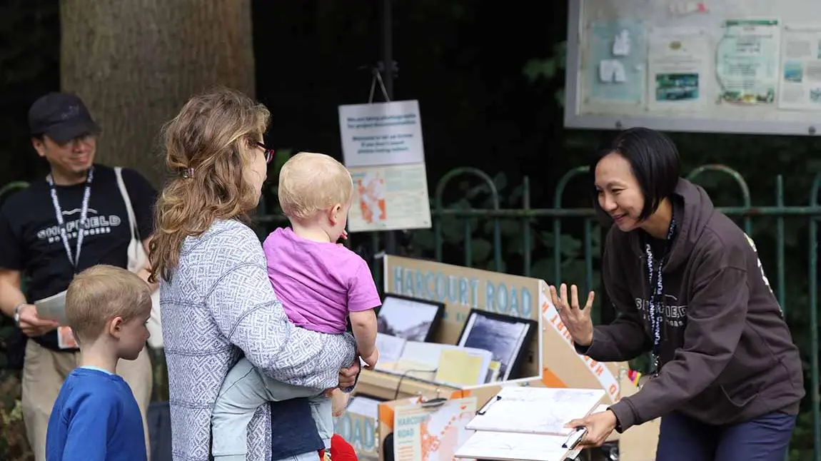 A woman stands outside in front of a bike-mounted display of pictures and other exhibits. She holds a clipboard and talks to a woman and two young children.