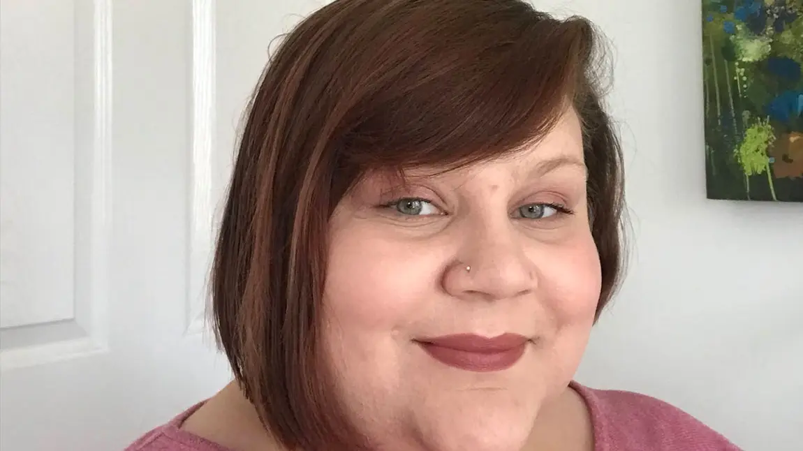 A selfie of a women with shoulder-length brown hair, standing against a white wall