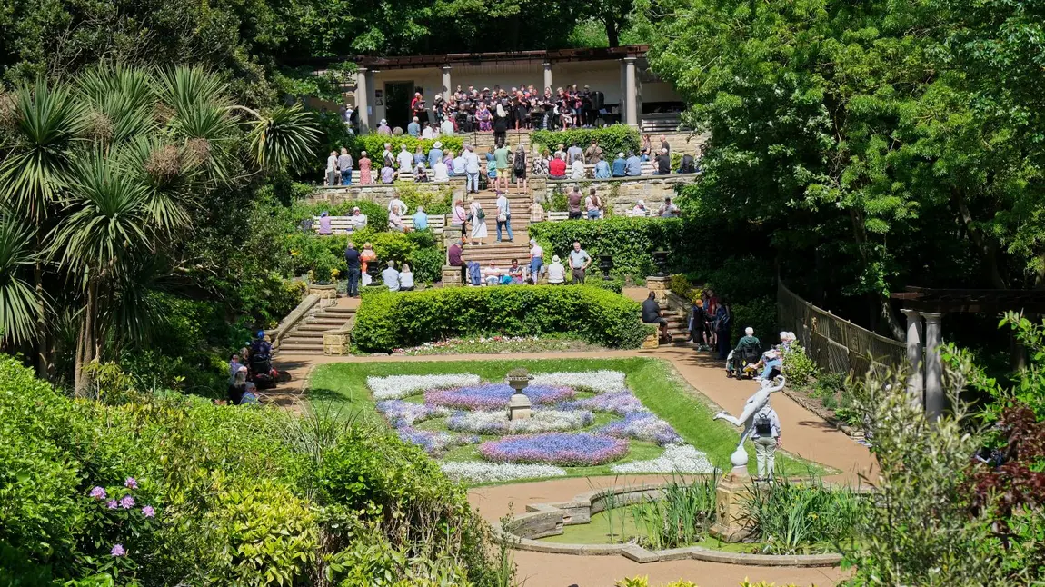 A view of landscaped gardens, featuring a fountain. A choir are stood above steps at the top of the gardens.
