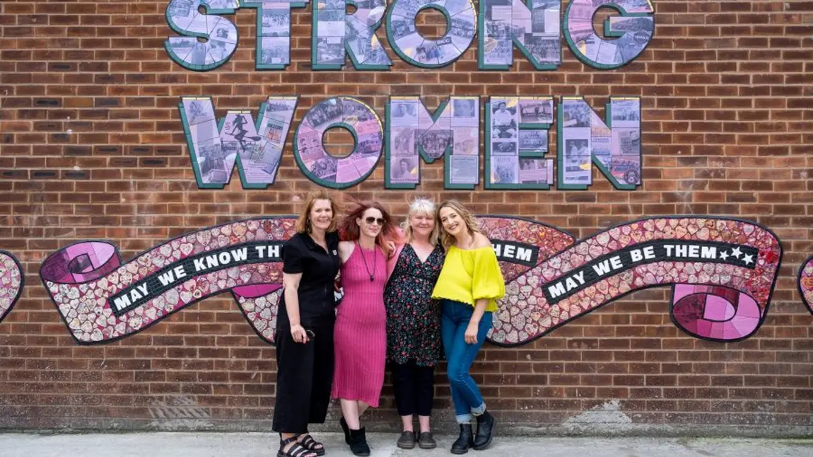 People standing in front of a ceramic mural that spells out the words Strong Women