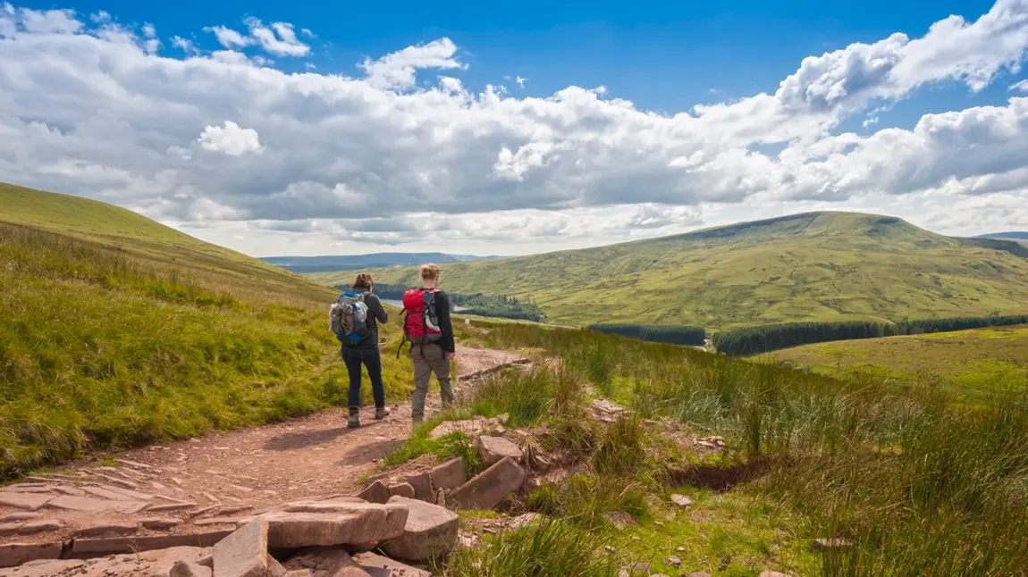 Two hikers walk away down a dirt track into an open, green expanse of the Brecon Beacons, on a sunny day.