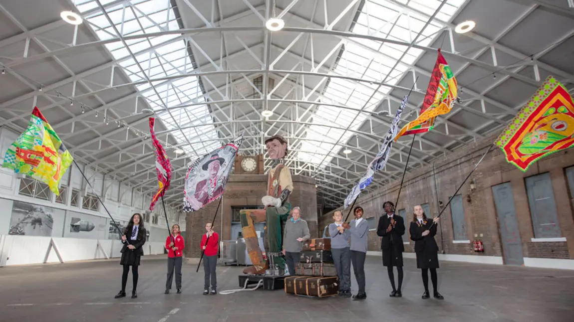 School children holding large flags stand in an empty station building accompanied by a giant puppet.