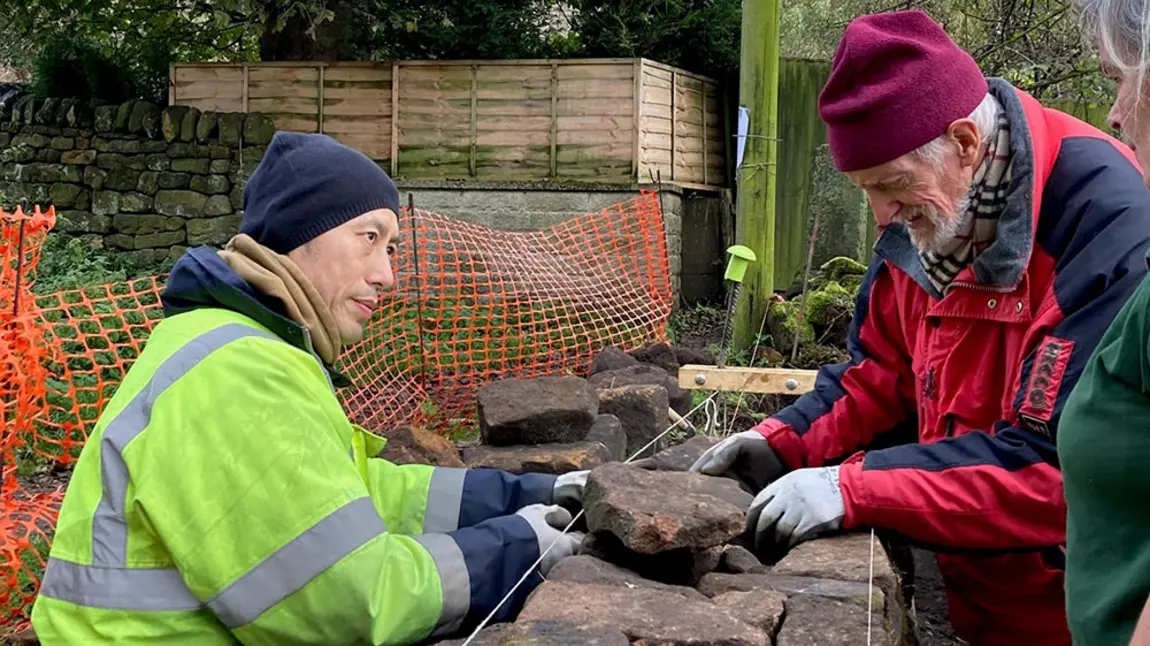 A person demonstrates dry stone wall construction techniques to two people in an outdoor setting