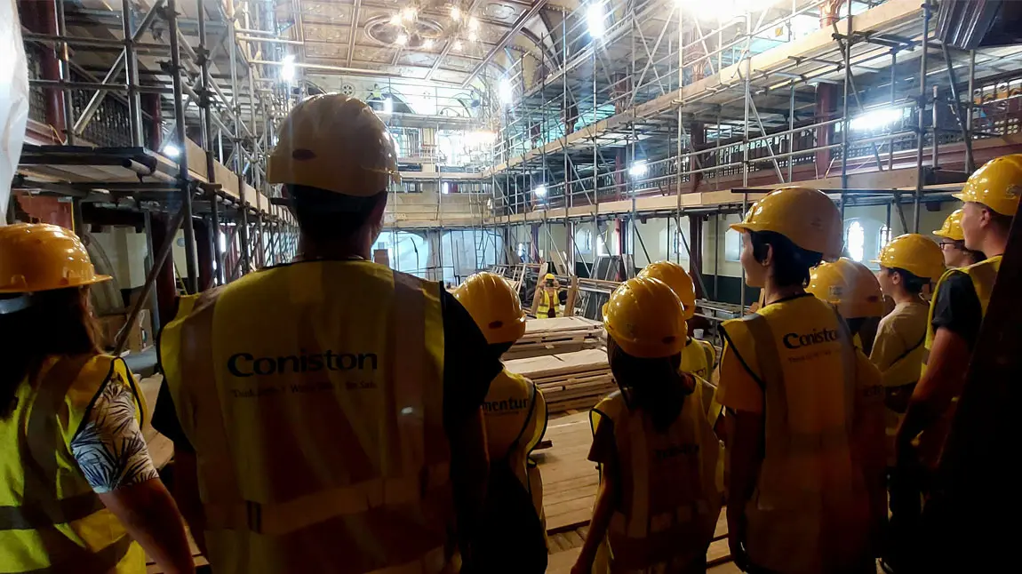 a group of people wearing hard hats and high-vis jackets stand inside the building looking at conservation works