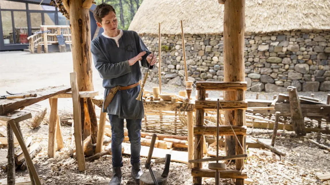 A person demonstrates Iron Age wood working techniques standing in a reconstructed Iron Age roundhouse