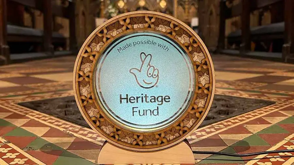 The National Lottery Heritage Fund acknowledgement stamp etched onto a circular glass plaque with a fretwork boarder. The plaque sits in front of a colourful tiled floor and an ironwork grate.