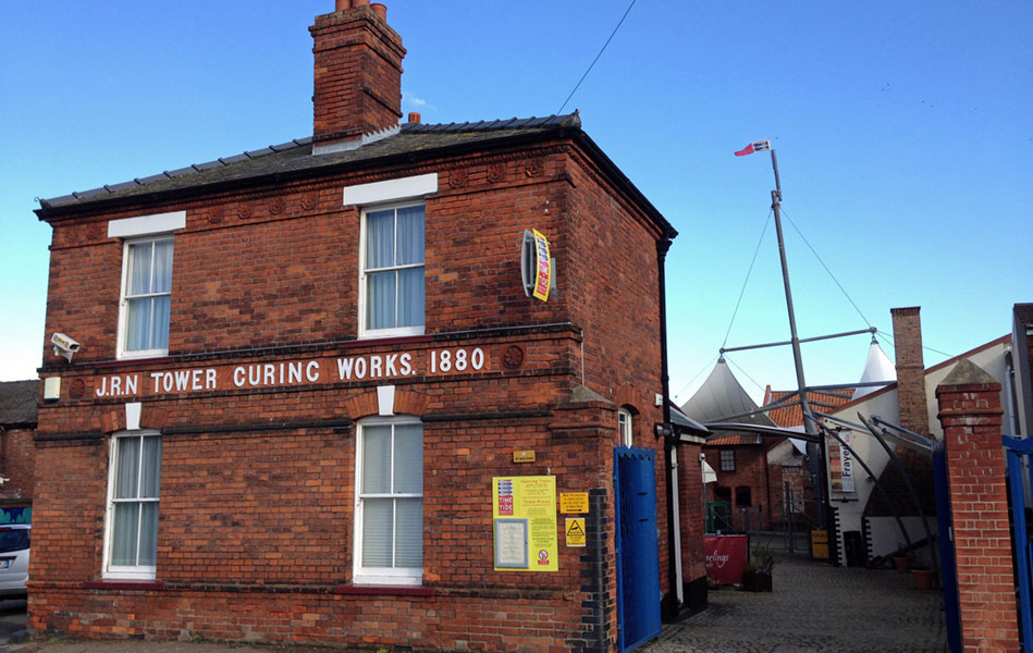 A red brick building with ‘J.R.N Tower Curing Works. 1880’ written on the front. A driveway with an open blue gate is on the right of the building.