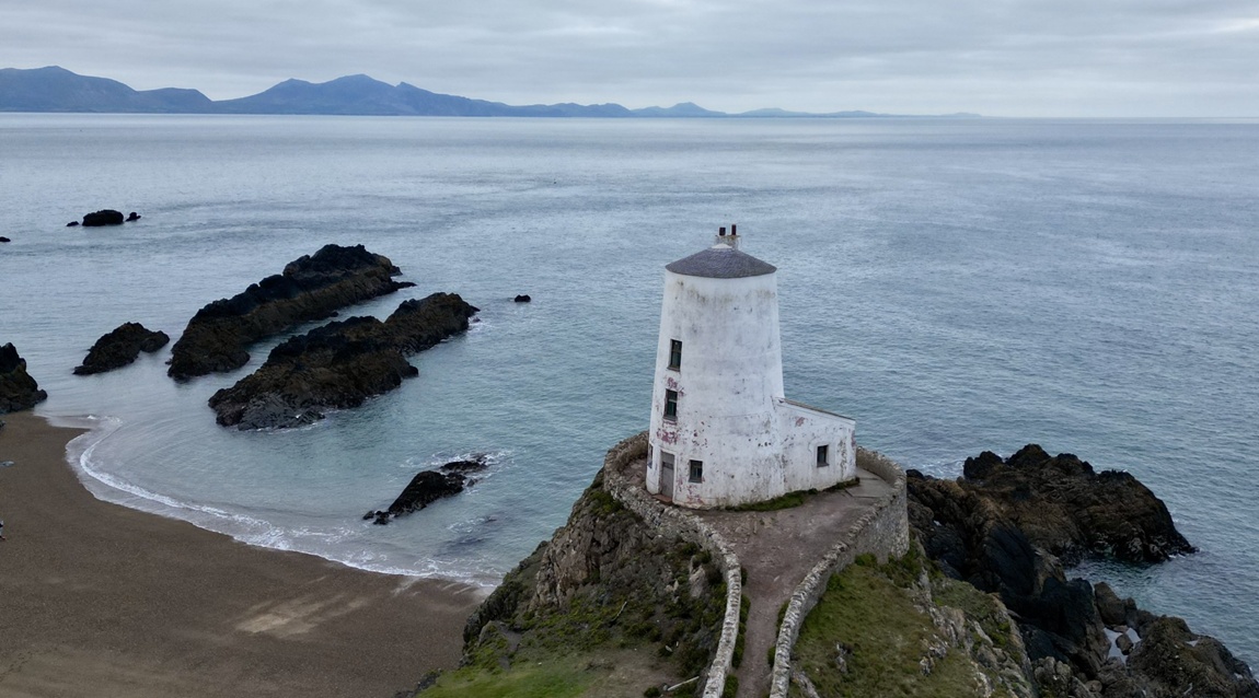 A lone white lighthouse sits atop a stack of rock on the craggy shoreline of Ynys Mon, looking out over the Menai Strait.
